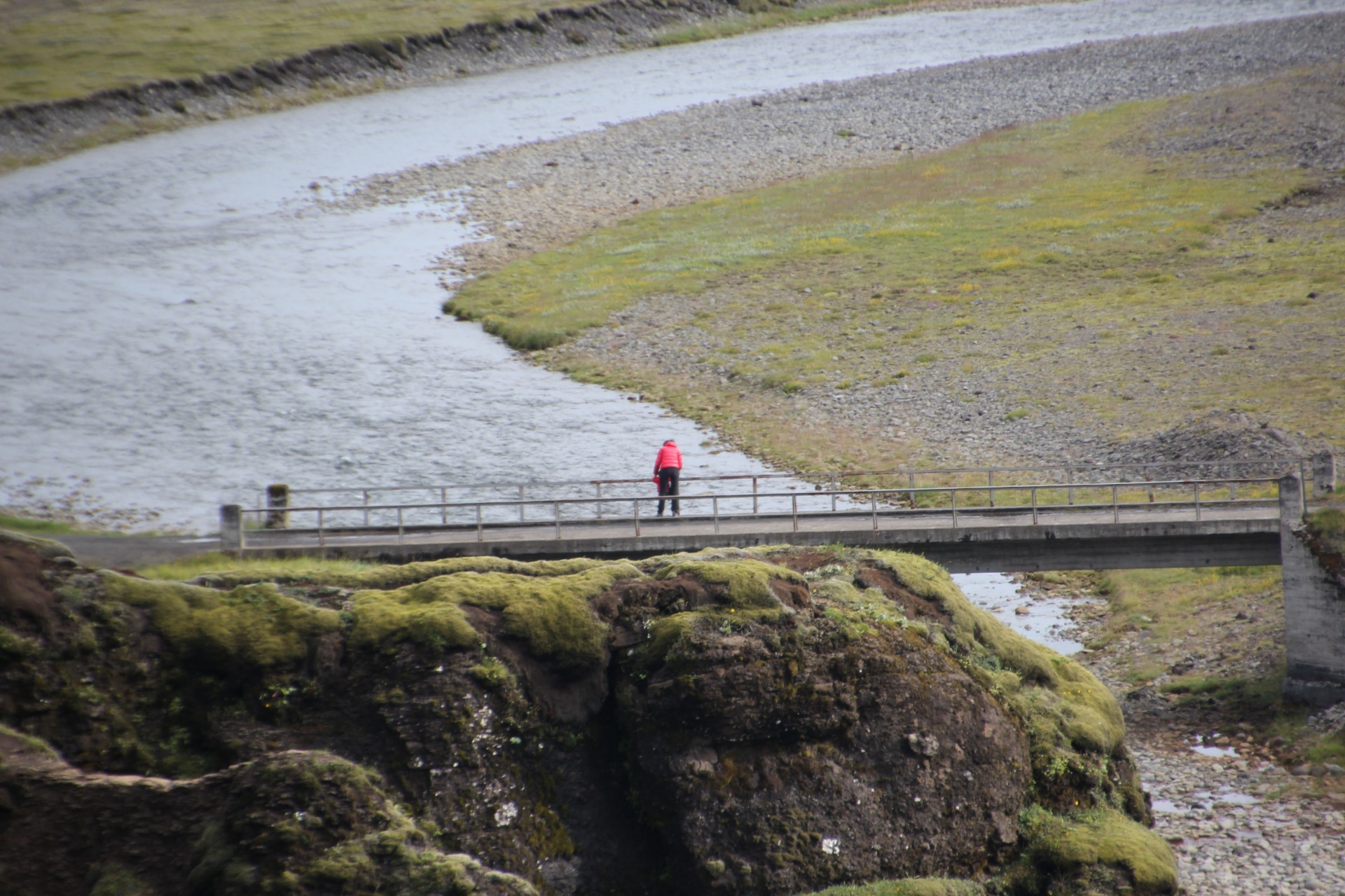 A lone figure in red on a small bridge over an Icelandic river